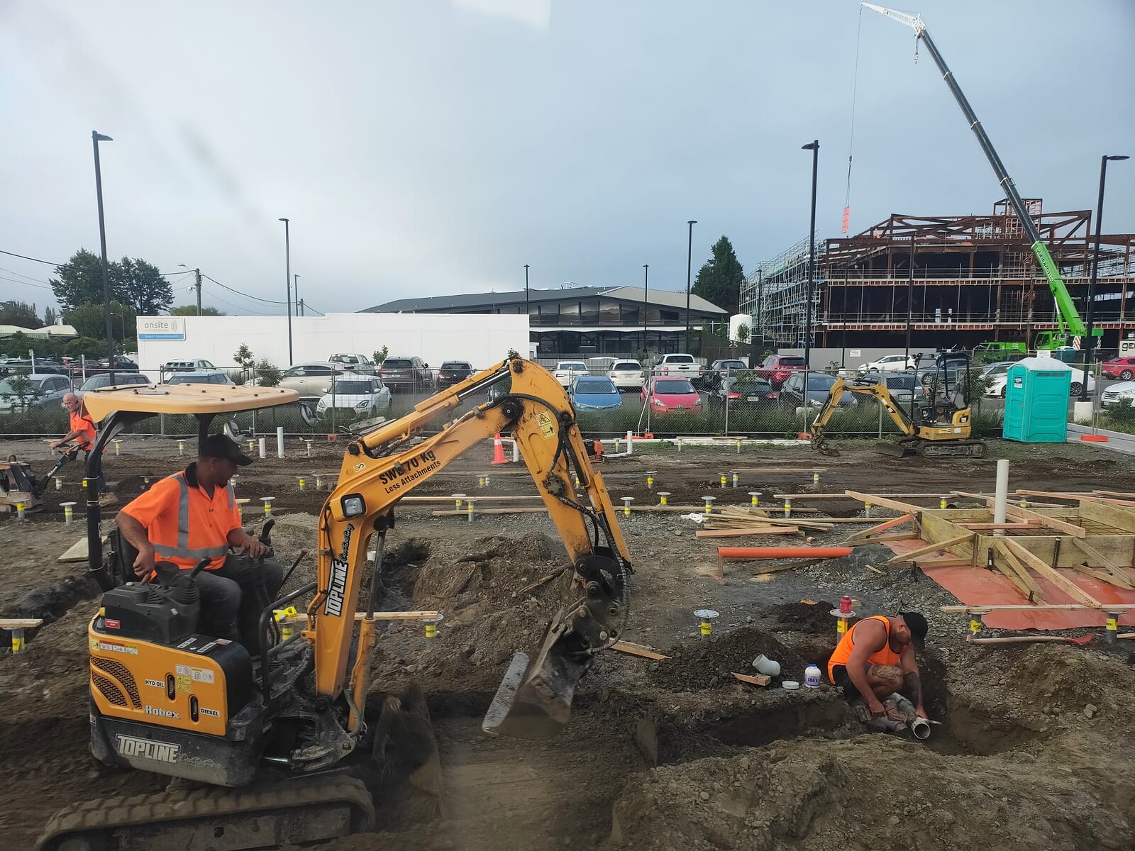 An excavator and ground crew setting out foundations and services across the site